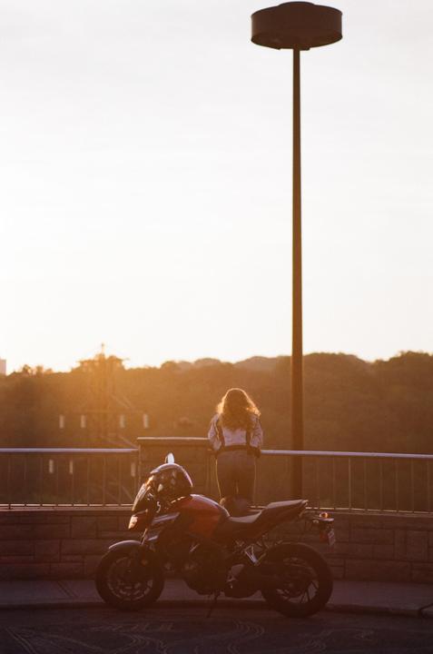 A woman stands on a bridge next to a motorcycle, looking out at a sunset landscape.