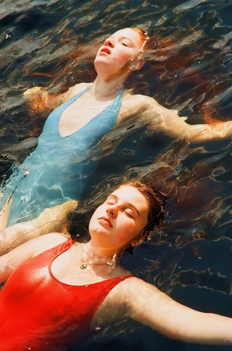 Two women float on their backs in dark water, one in a red swimsuit and the other in a blue swimsuit.