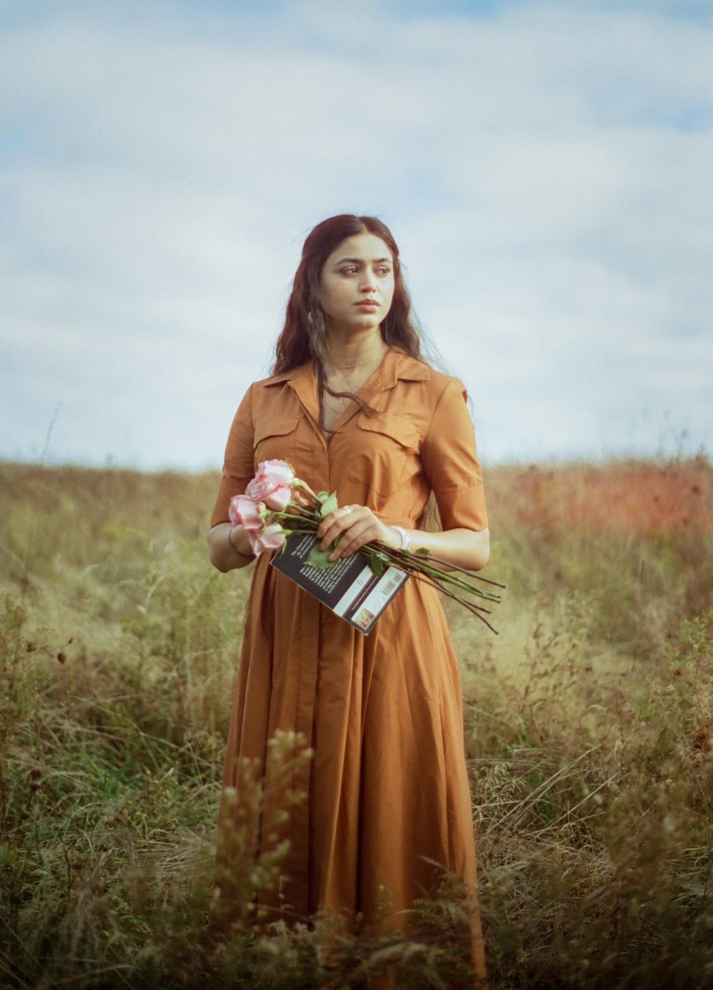 A woman in a brown dress stands in a field holding a book and a bouquet of pink roses.