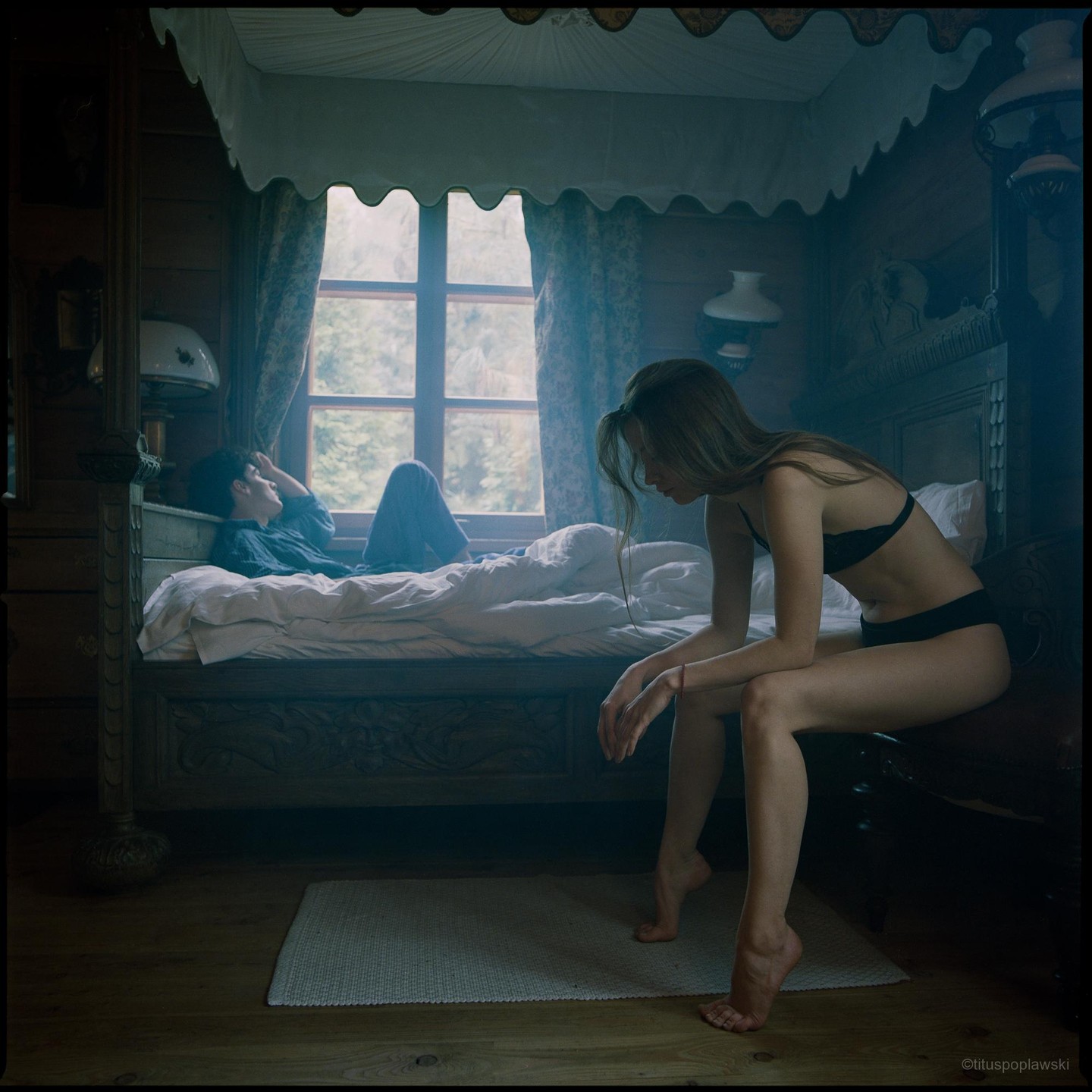 A woman in lingerie sits on a chair while a man lies on a bed looking out a window in a rustic room.