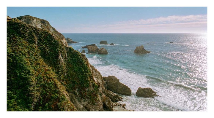 A steep, green, rocky cliff overlooks a sparkling ocean with waves crashing on the shore and several large rocks protruding from the water.