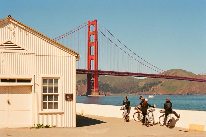 A group of people on bicycles are gathered near a white building with the Golden Gate Bridge in the background.