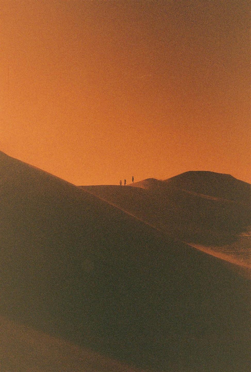 Three people stand silhouetted on a sand dune against an orange sky.