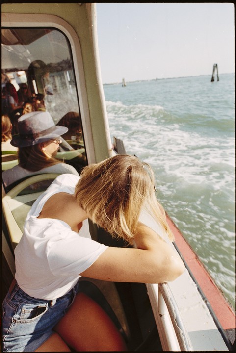 A woman with blonde hair leans out of a boat, looking at the water, while another woman in a hat sits behind her.