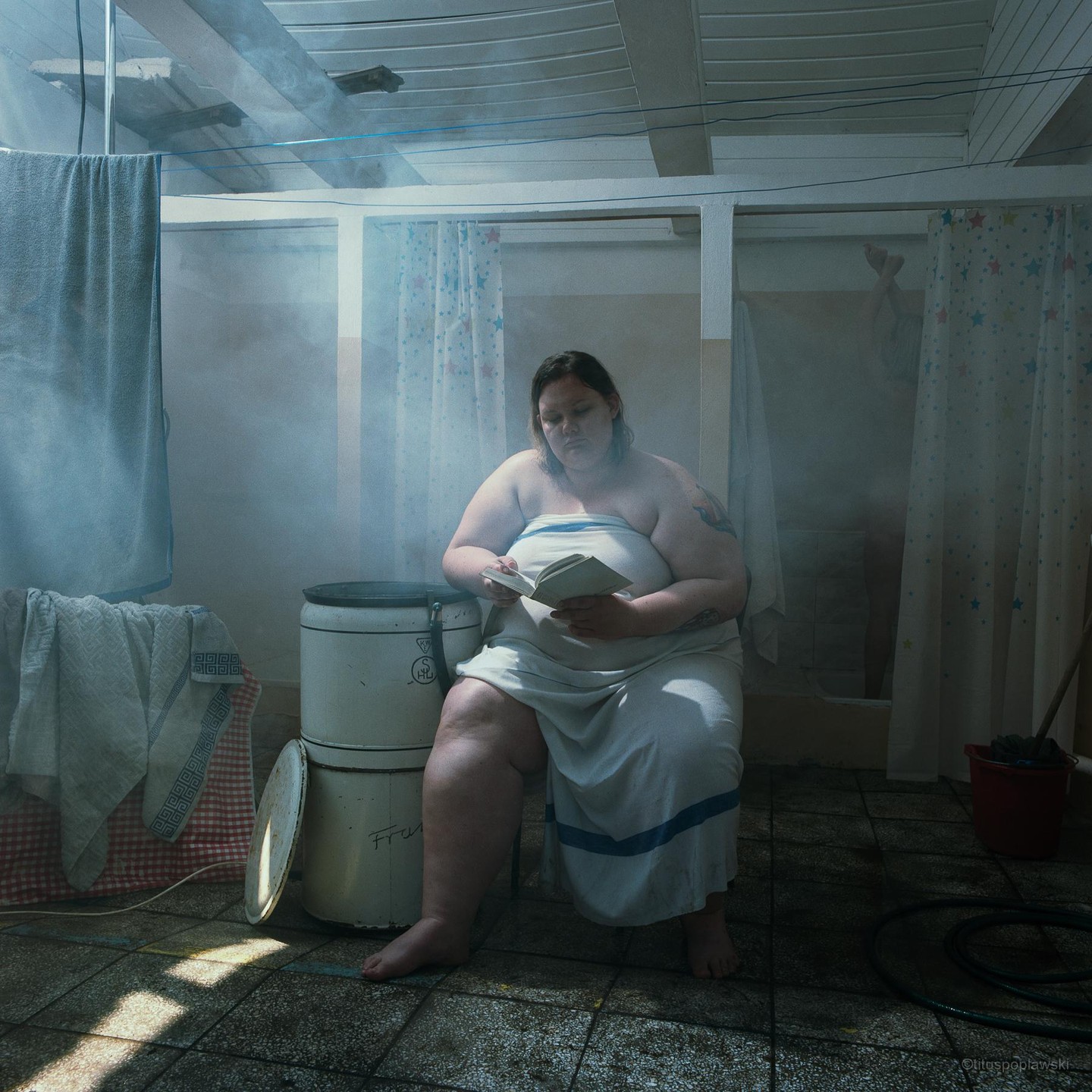 A woman wrapped in a towel sits on a stool reading a book in a steamy room with shower curtains.
