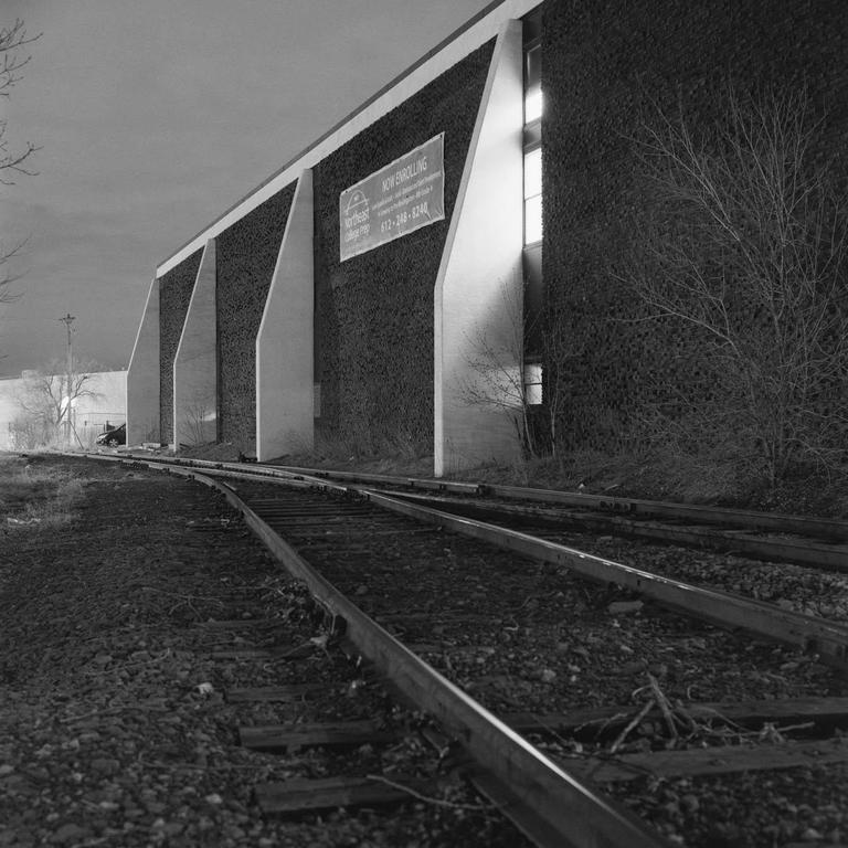 A building with a "Now Enrolling" sign stands next to railroad tracks at night.