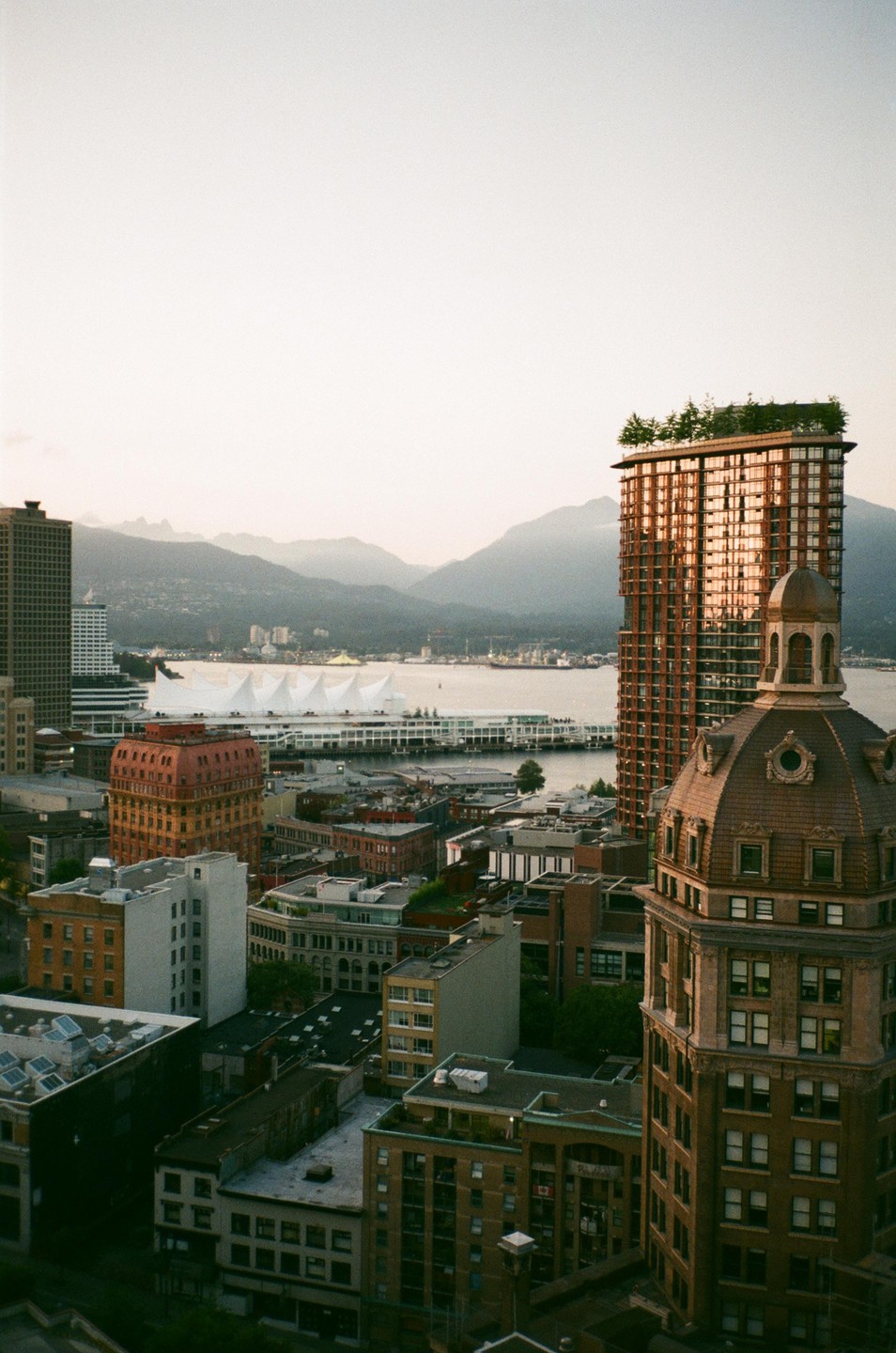 An aerial view of downtown Vancouver shows various buildings, a waterfront convention center, and mountains in the background under a hazy sky.