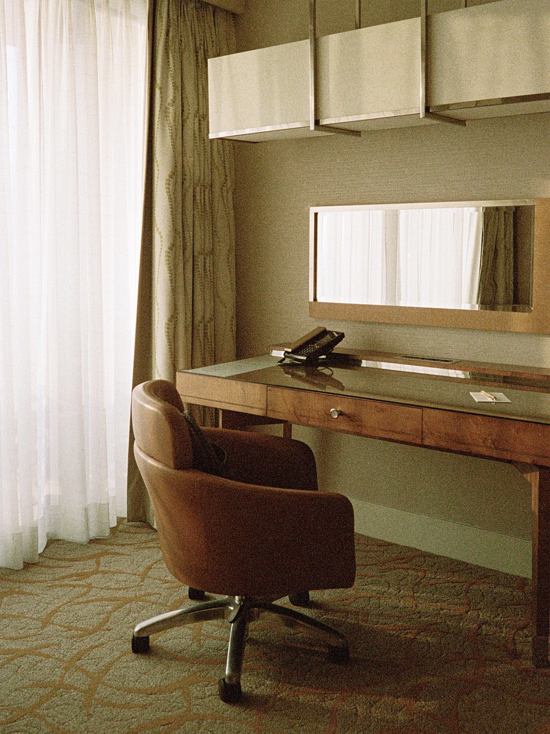 A brown office chair sits in front of a wooden desk with a mirror and telephone, next to a window with sheer white curtains.