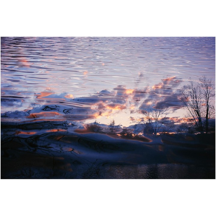 The sunset sky with clouds and bare trees is reflected in the rippling water of a lake.