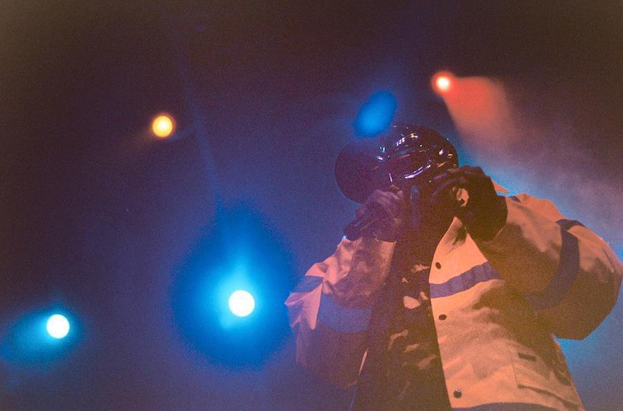 A man wearing a helmet and jacket performs on stage under colorful lights.