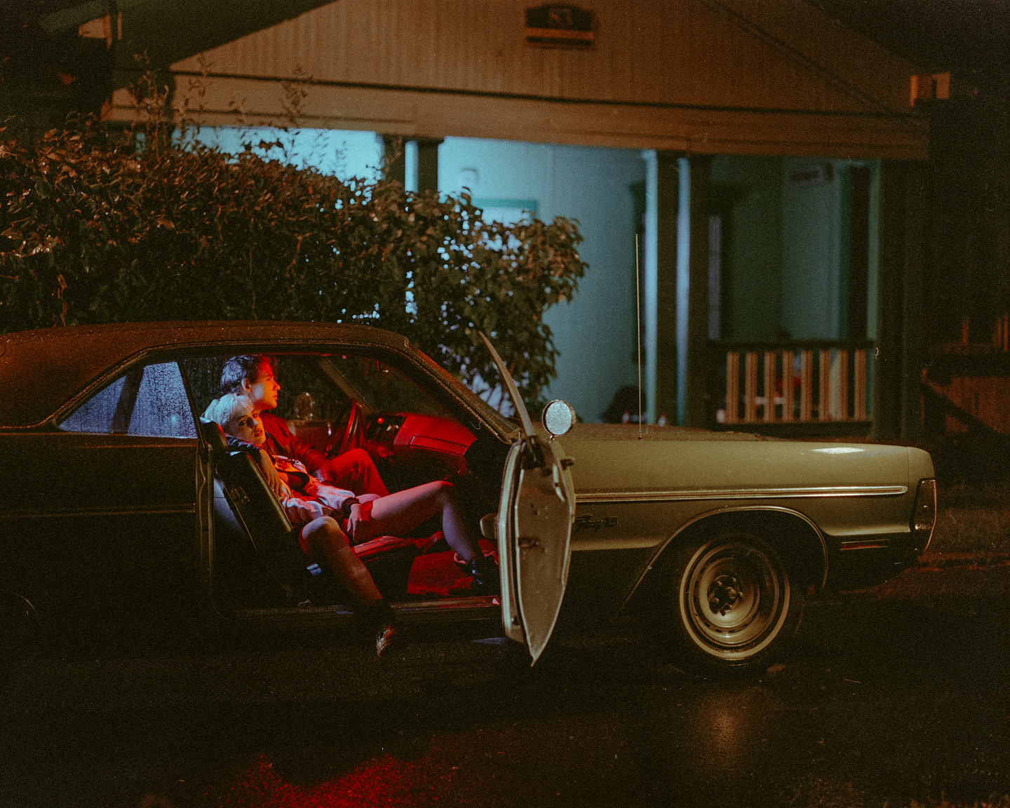 A couple sits in a car with an open door at night, illuminated by red light.