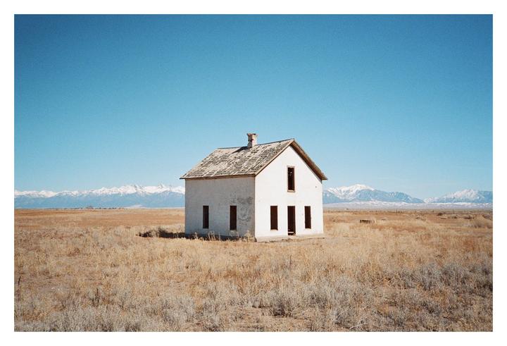 A small, white, abandoned house stands alone in a dry, grassy field with snow-capped mountains in the distance under a clear blue sky.