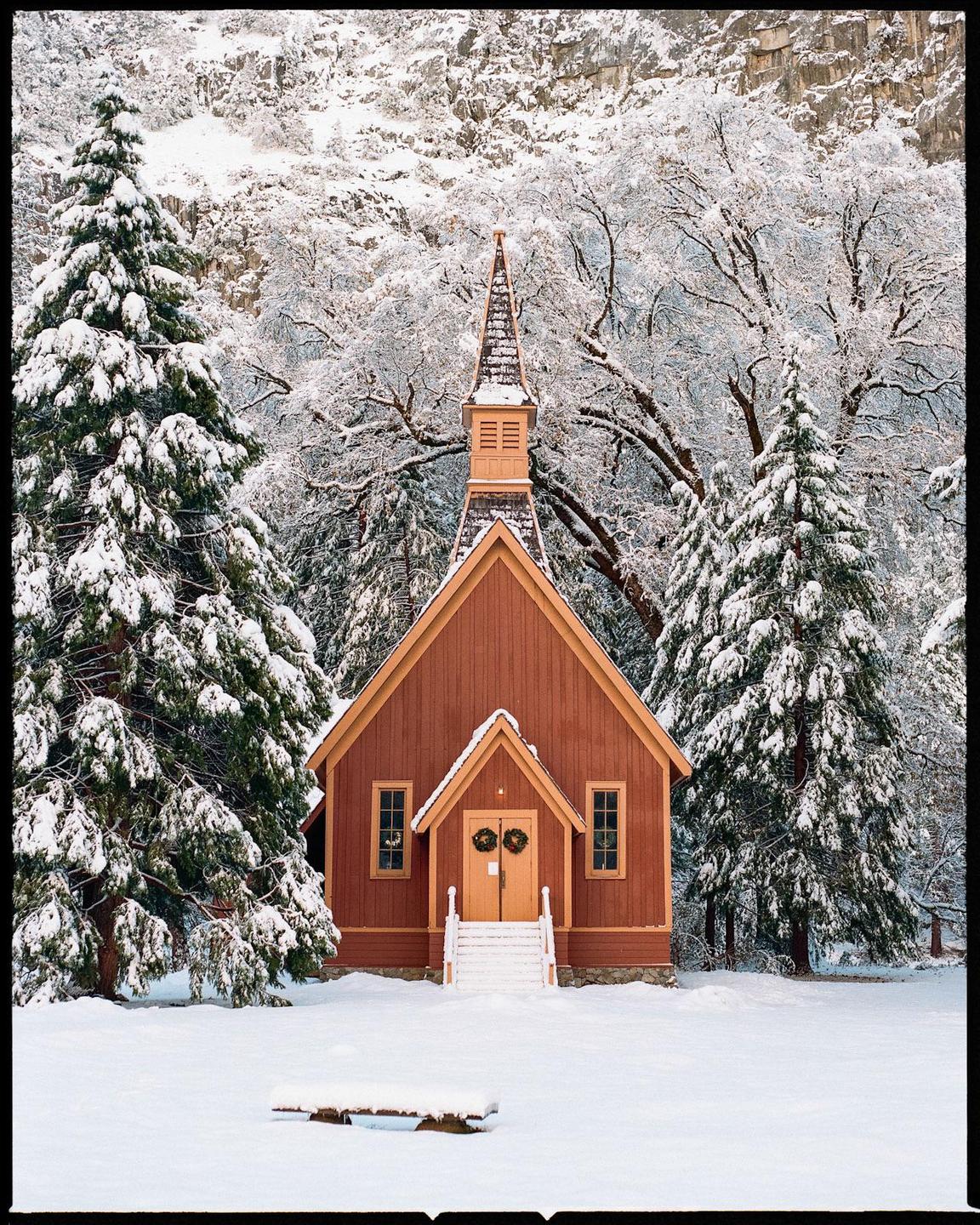 A small red chapel with a steeple stands in a snowy landscape surrounded by snow-covered trees.