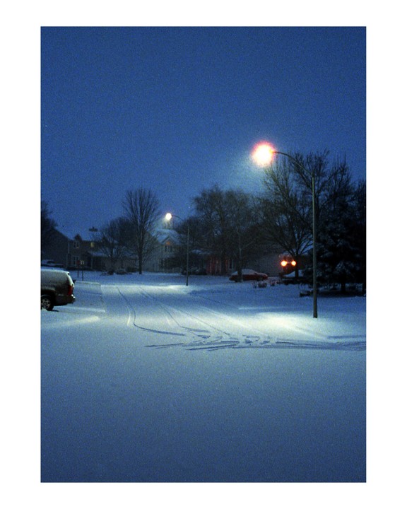 A snow-covered street is illuminated by streetlights at dawn.