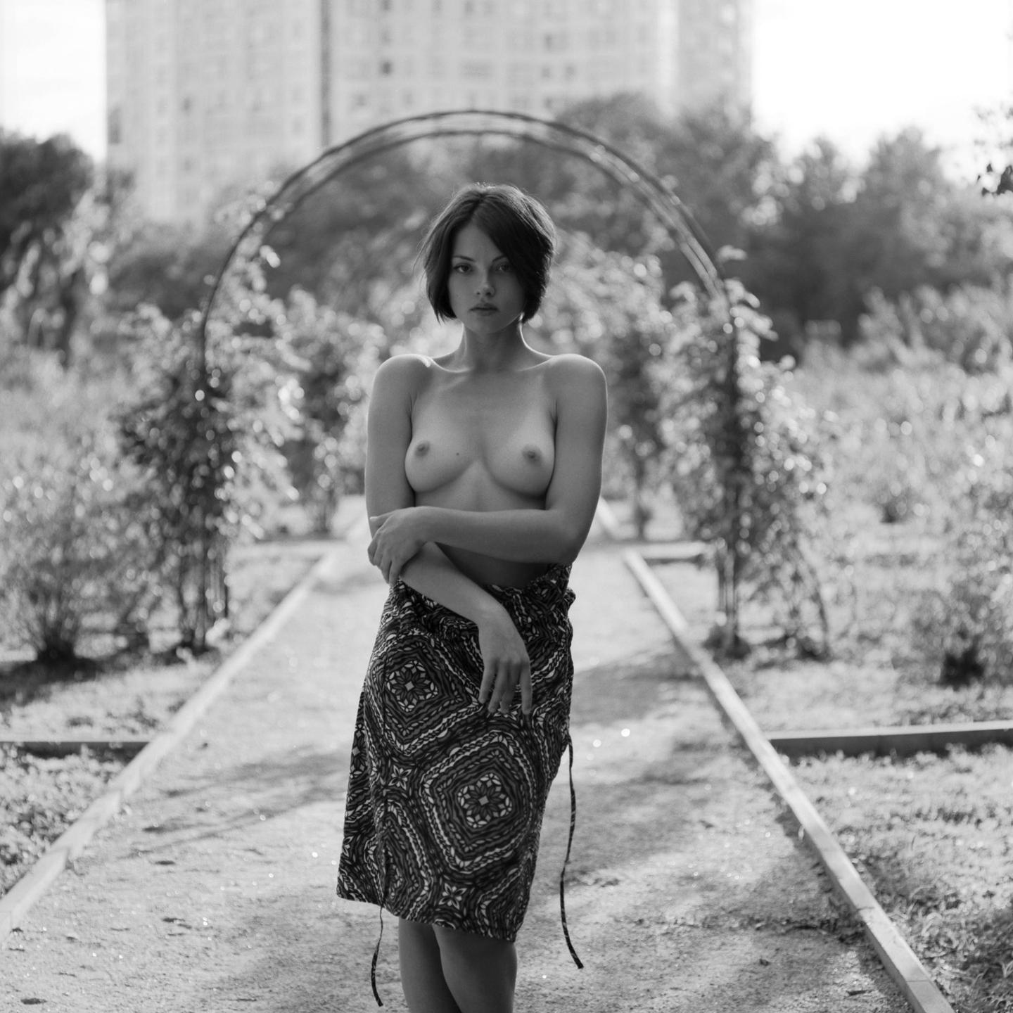 A topless woman with a patterned skirt stands on a path under an archway in a garden.