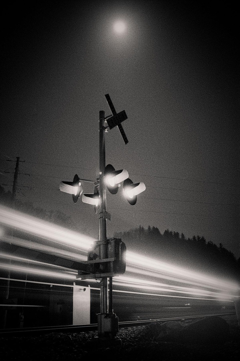 A railroad crossing signal stands under a bright moon with light trails from a passing train.