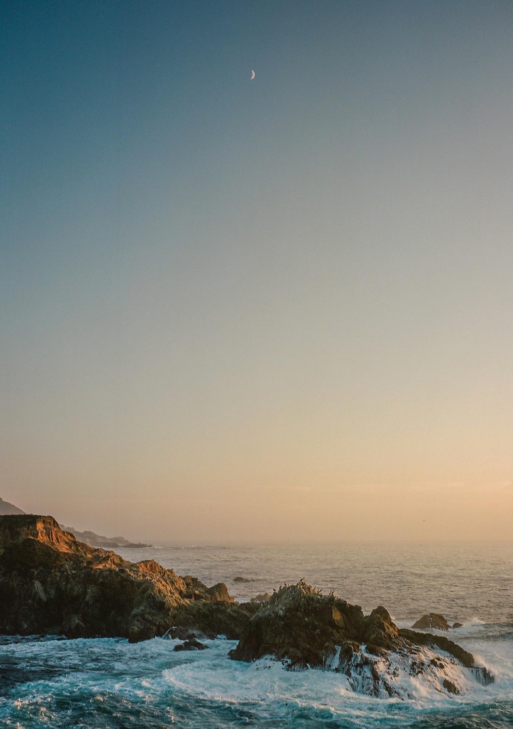 A crescent moon hangs in the sky above a rocky coastline with waves crashing against the shore at dusk.