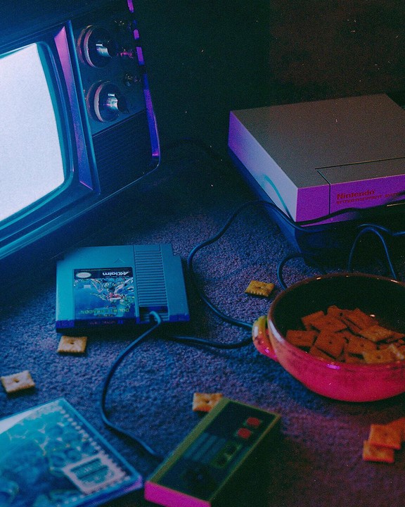 A television, a Nintendo console, a video game cartridge, a controller, and a bowl of crackers are arranged on a carpet.