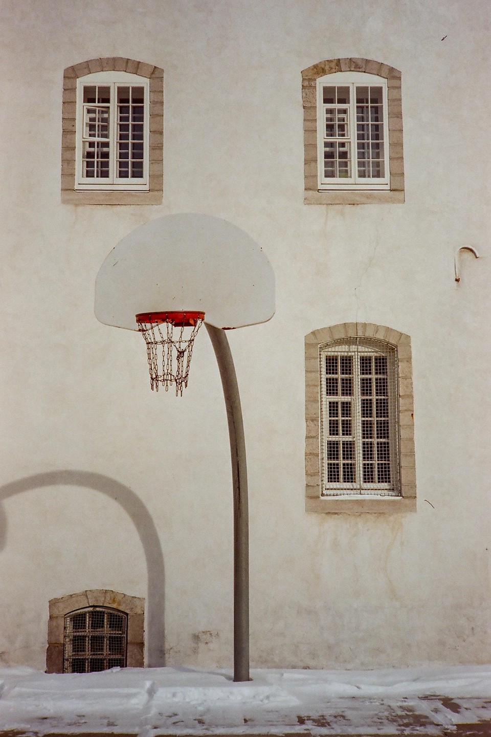 A basketball hoop with a chain net stands in front of a white building with several windows, with snow on the ground.