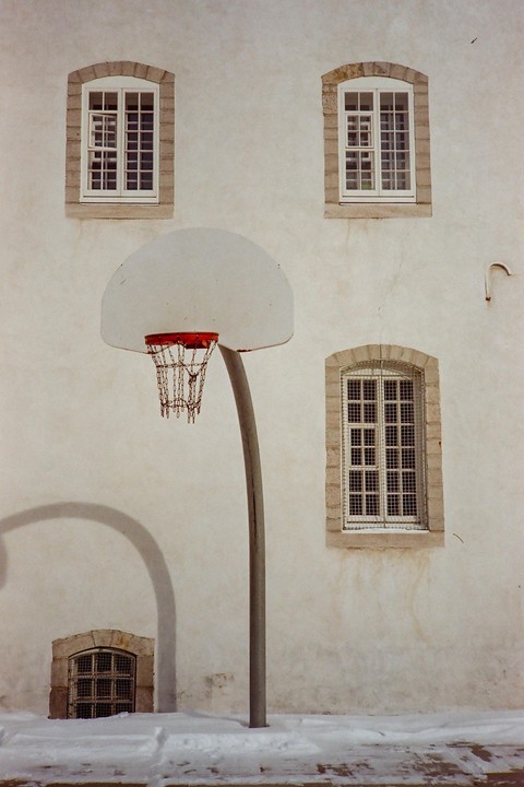 A basketball hoop with a chain net stands in front of a white building with several windows, with snow on the ground.