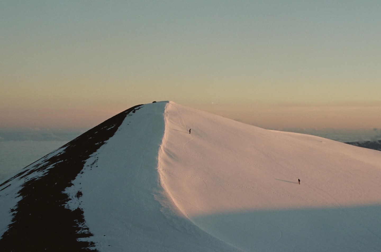 Two people walk on a snow-covered mountain ridge at sunset, with clouds visible in the distance.