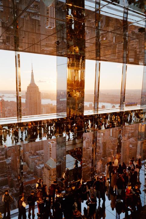 A crowd of people stands on a mirrored floor and looks out at the New York City skyline with the Empire State Building visible in the distance.