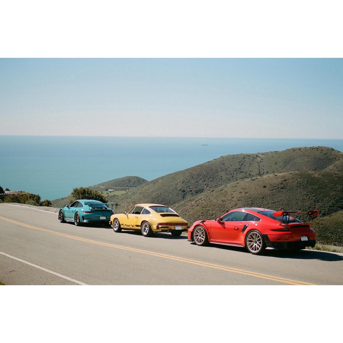 Three sports cars are parked on the side of a winding road overlooking the ocean and hills.