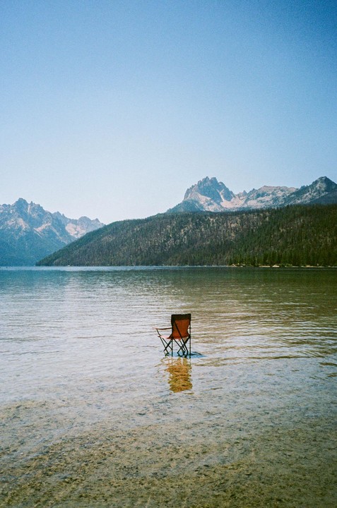 An orange camping chair sits in clear lake water with mountains and a forest in the background under a clear sky.