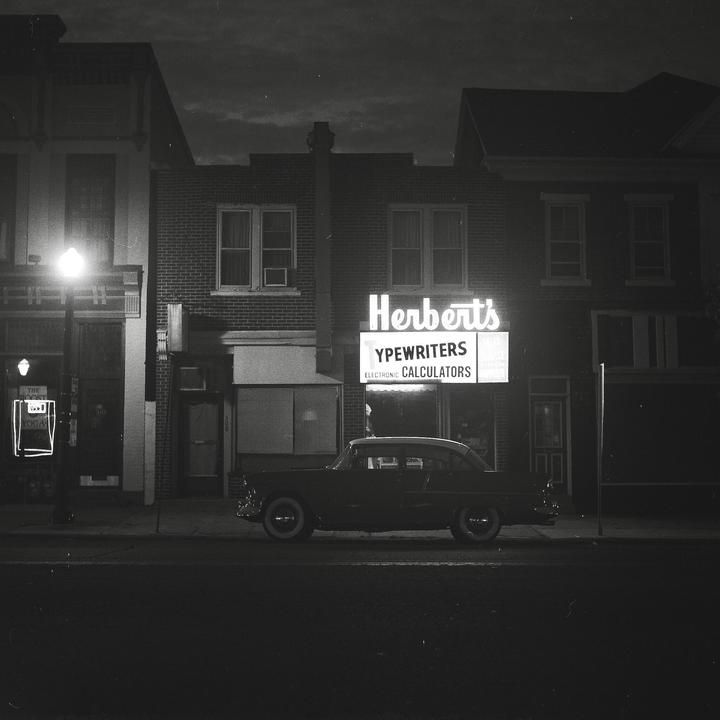 A vintage car is parked on a street in front of a building with a neon sign for Herbert's Typewriters and Calculators.