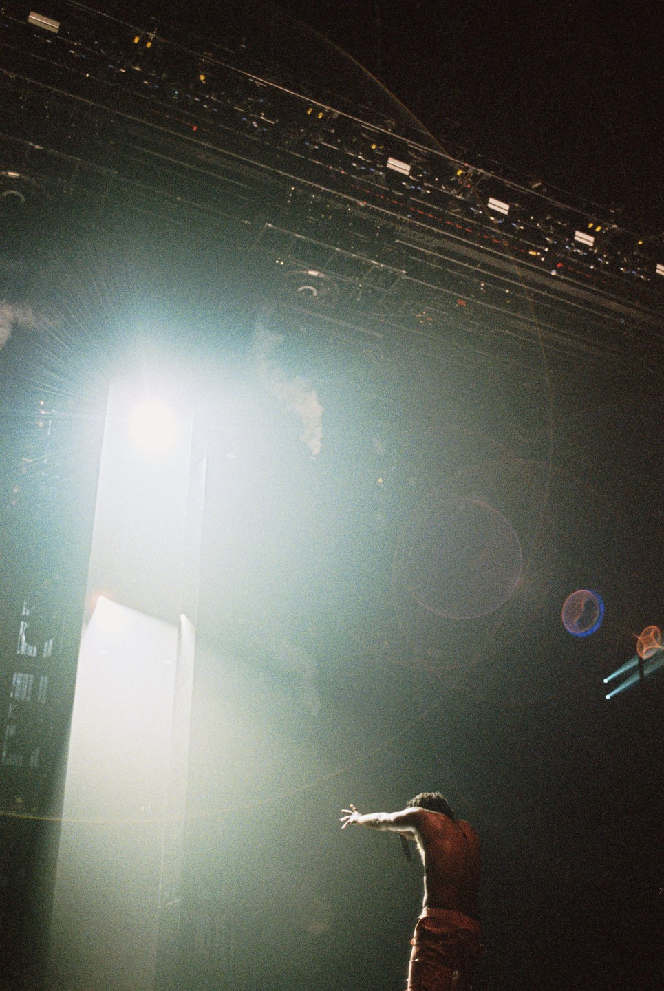 A shirtless man with dreadlocks stands on a stage with his arms outstretched towards a bright light.