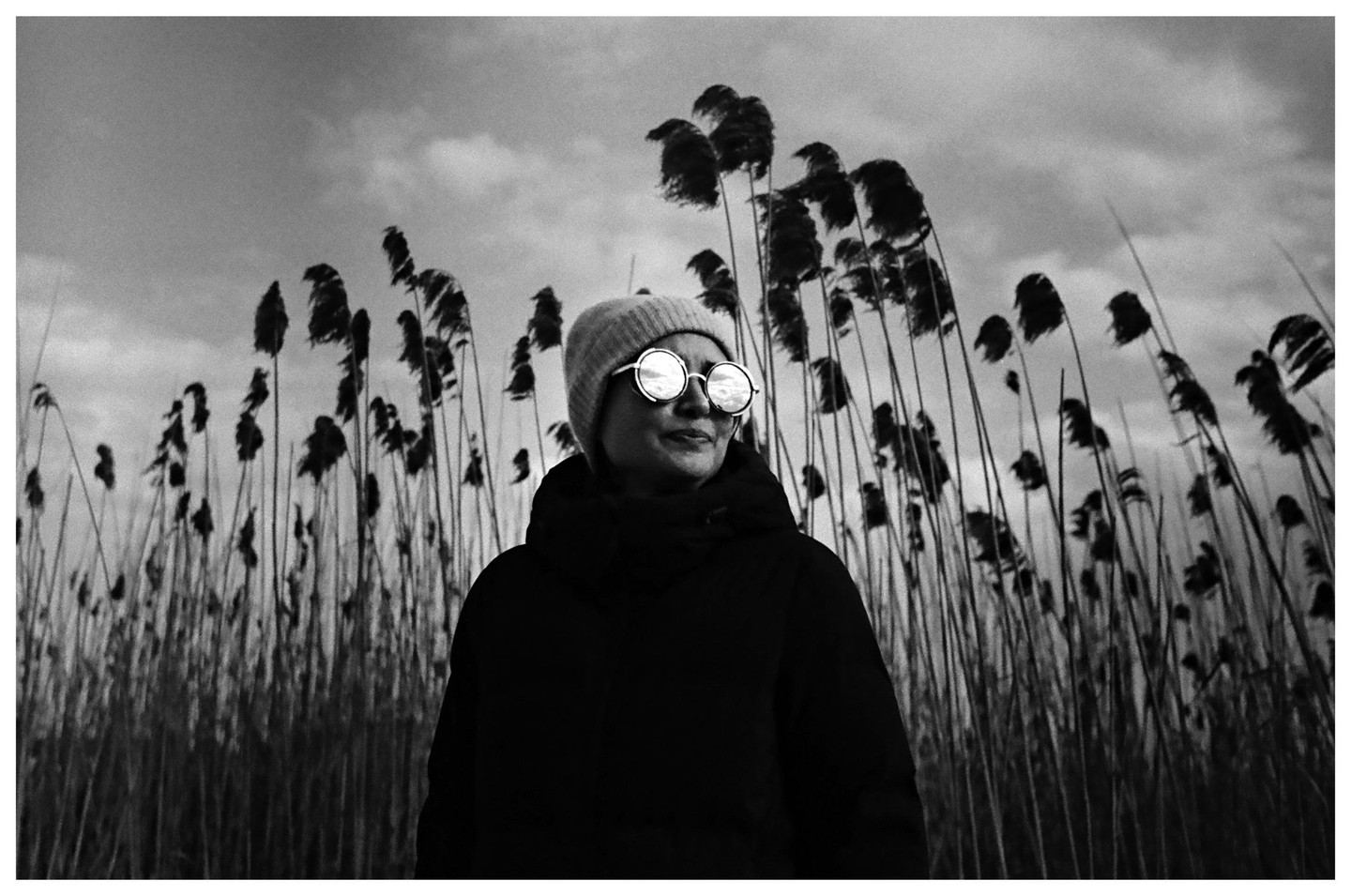 A woman wearing a beanie and round sunglasses stands in front of tall reeds under a cloudy sky.