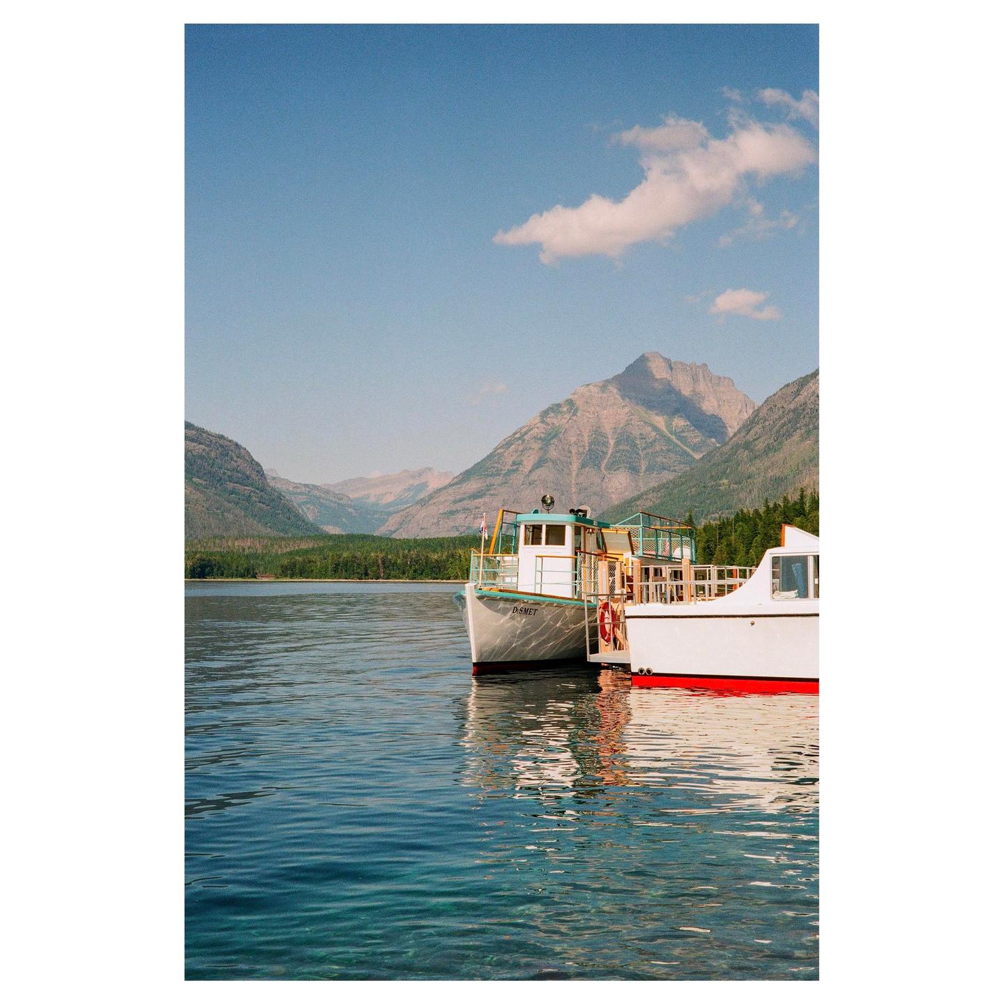 Two boats are docked on a clear lake with mountains and a forest in the background under a blue sky.