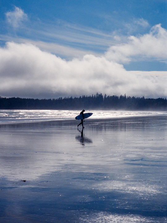 A surfer walks along a wet beach with a surfboard under a cloudy sky.
