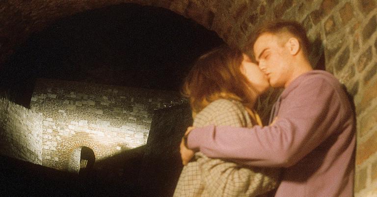 A couple kisses in a dark stone tunnel with a brick wall visible in the background.