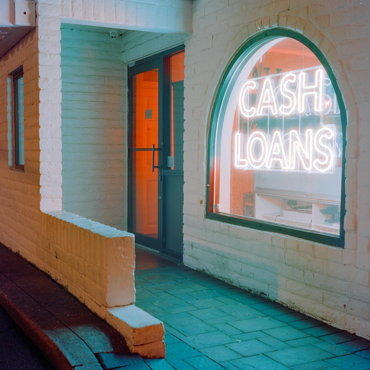 A neon sign for "CASH LOANS" glows in an arched window next to a doorway with an orange-lit interior.