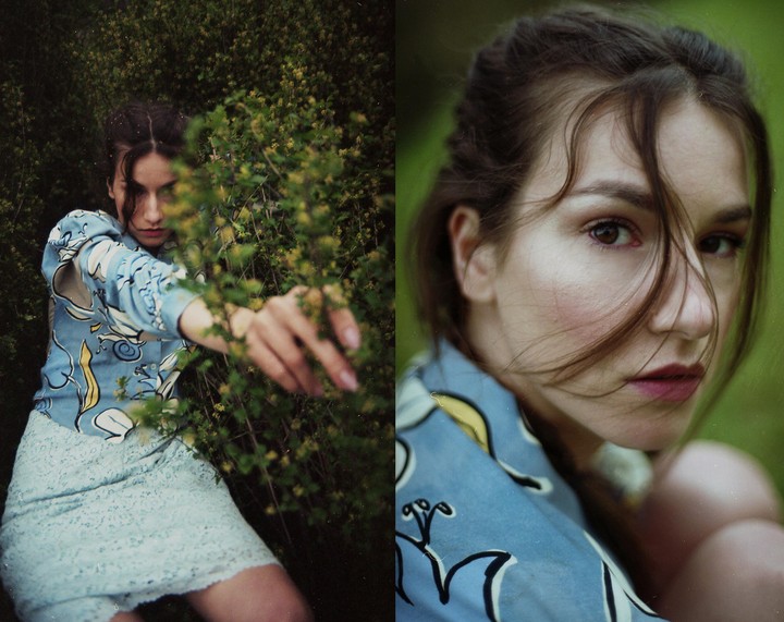 A woman in a blue patterned shirt and light blue skirt is partially obscured by green foliage.