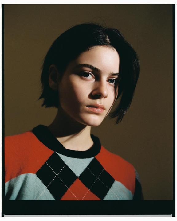 A woman with short dark hair wears an argyle sweater and looks towards the viewer.
