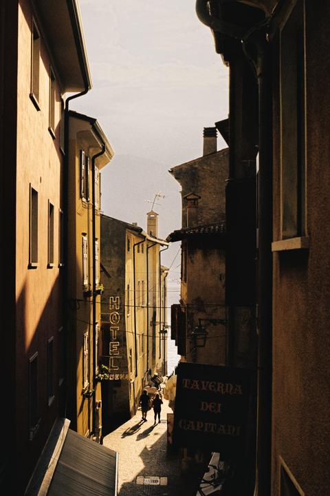 Two people walk down a narrow street between tall buildings with a view of water at the end.