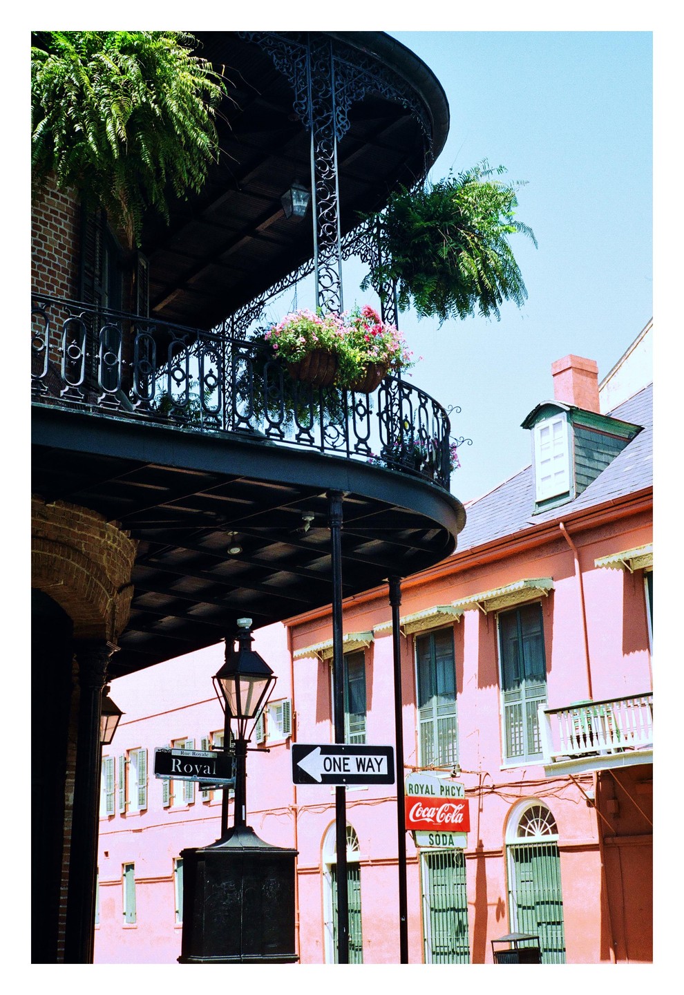 A black wrought iron balcony with hanging plants extends over a street with a pink building and a street sign.