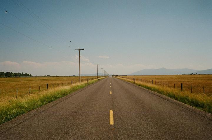 A long, straight road with a yellow dashed line down the middle stretches into the distance, flanked by fields of golden grass and utility poles.