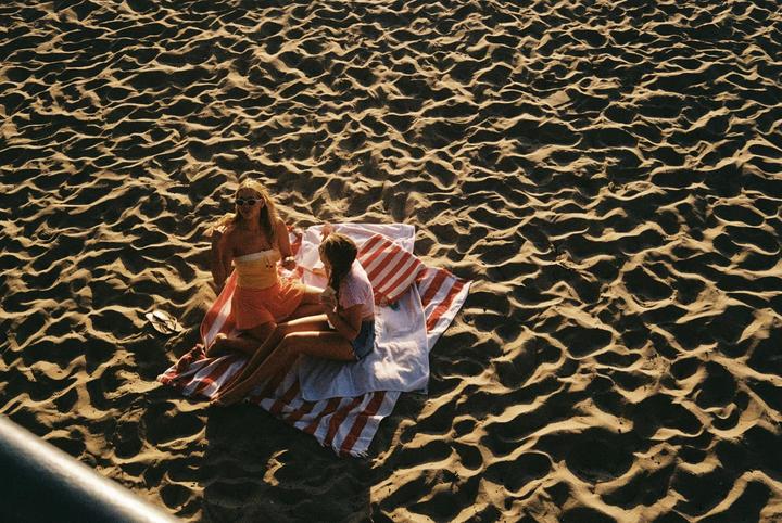 Two women sit on a striped towel on a sandy beach.