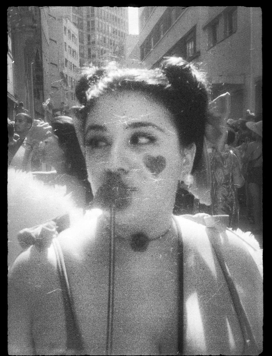 A woman with two buns in her hair, a heart painted on her cheek, and a fake mustache on a stick is looking to the side.