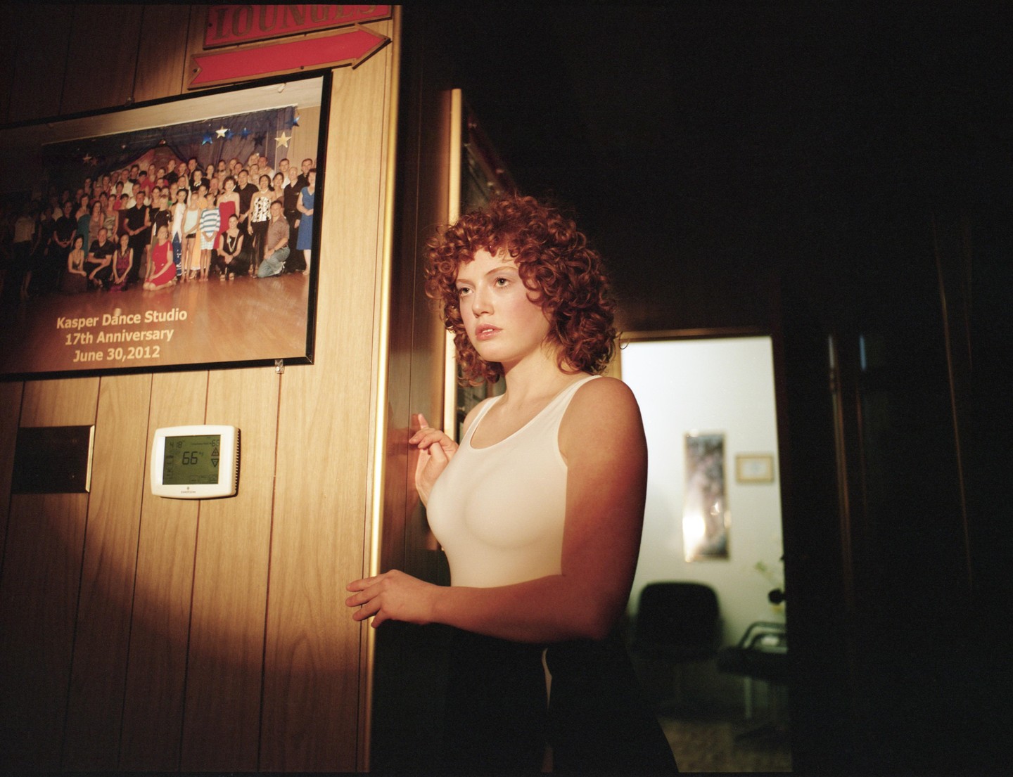 A woman with curly red hair stands next to a wooden wall with a framed group photo and a thermostat.