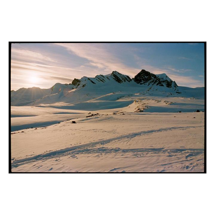 A snowy mountain range is illuminated by the setting sun, casting long shadows across the landscape.