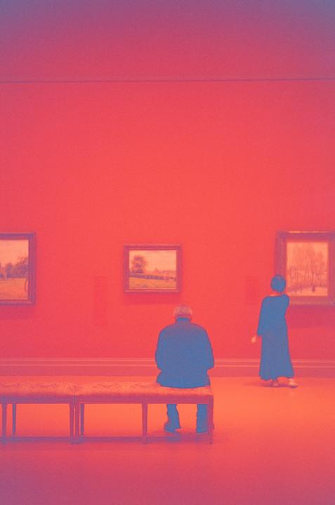 A man sits on a bench while a woman stands in an art gallery with red walls and paintings.