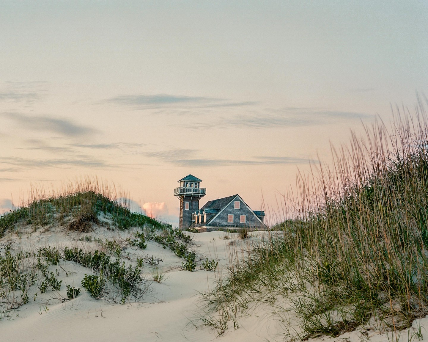A house with a tower is nestled among sand dunes with tall grass under a soft, cloudy sky.