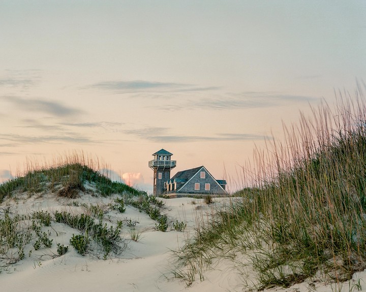 A house with a tower is nestled among sand dunes with tall grass under a soft, cloudy sky.