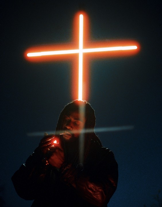 A man with dreadlocks lights a cigarette under a glowing orange cross.