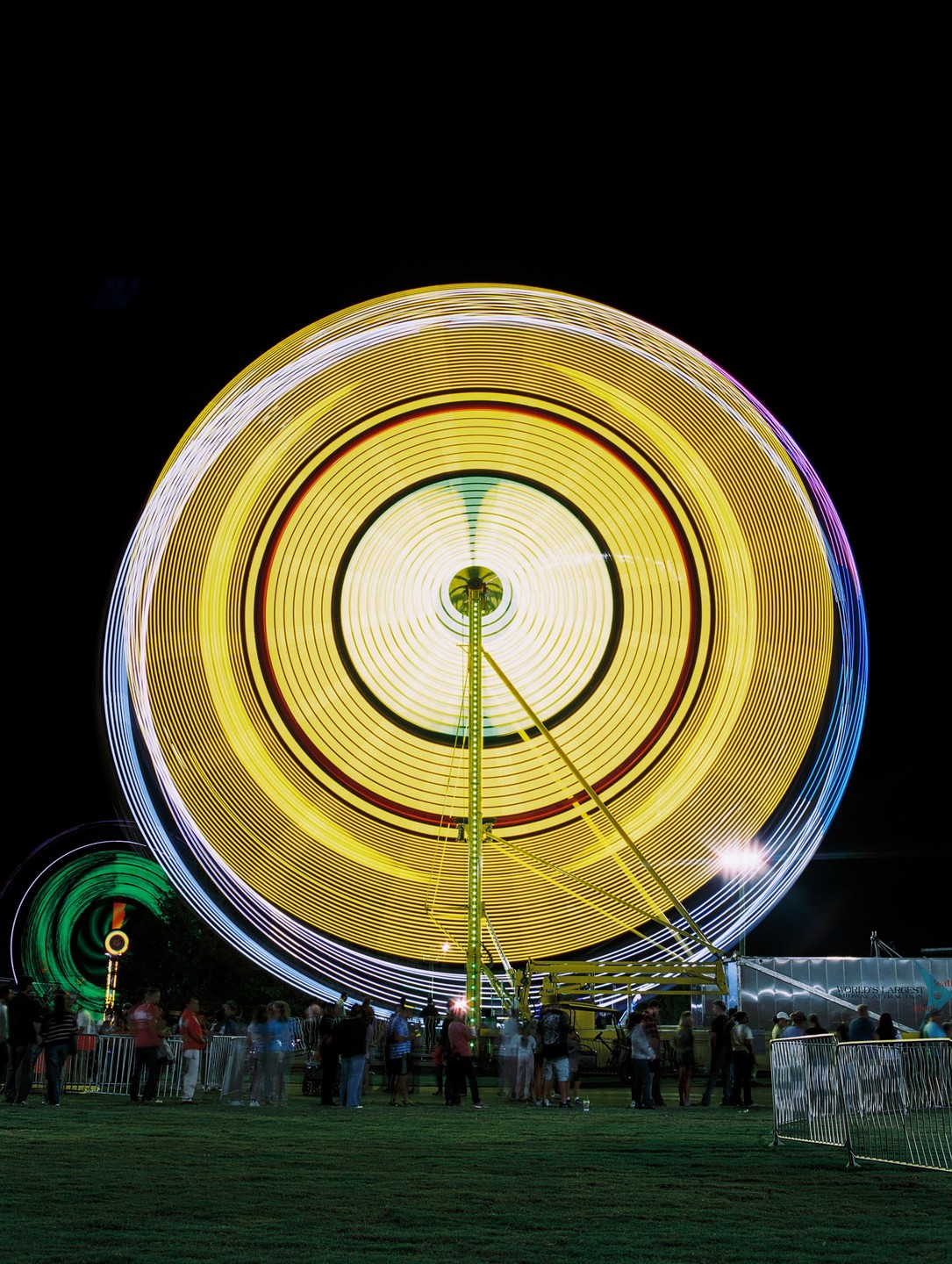 A long exposure shot captures the light trails of a spinning carnival ride at night, with people gathered on the grass below.