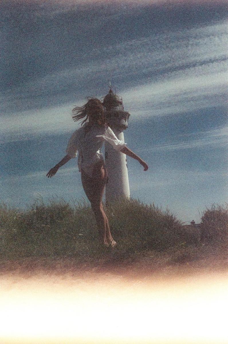 A woman with windblown hair stands in front of a lighthouse on a grassy hill under a cloudy sky.